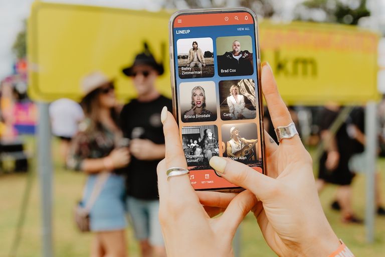 Person holding a smartphone displaying the Strummingbird music festival app lineup at an outdoor event.